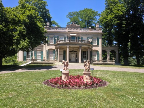 A grand historic mansion surrounded by lush greenery and flower beds, featuring two statues in the foreground.