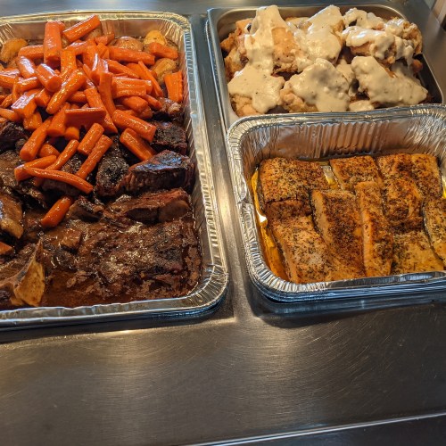 A tray of roasted carrots and beef, alongside a tray of biscuits with gravy and seasoned salmon fillets.
