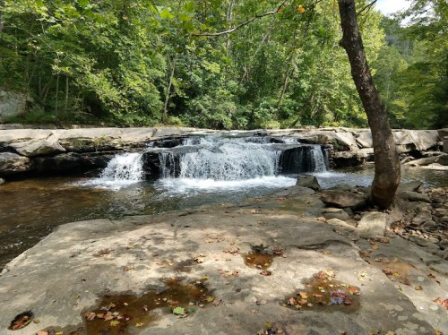 A serene waterfall cascades over rocks, surrounded by lush green trees and a peaceful riverbank.