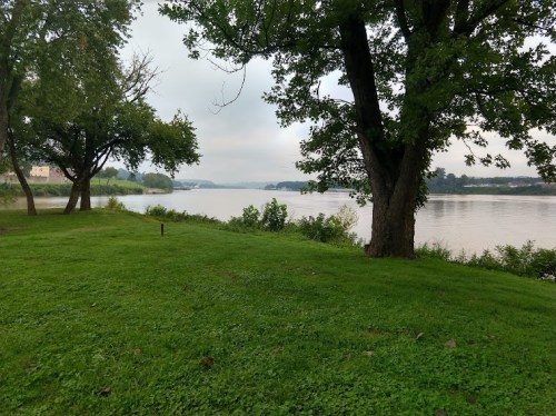 A serene riverside scene with green grass, trees, and a calm waterway under a cloudy sky.