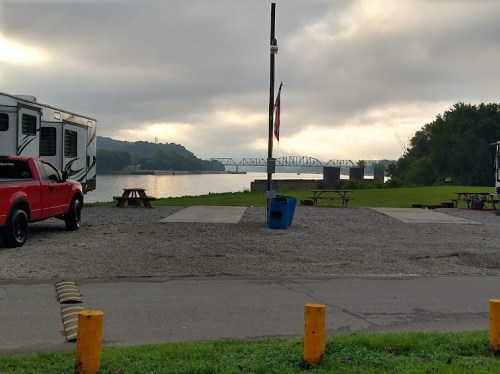 A serene riverside view with an RV, picnic area, and a bridge in the background under a cloudy sky.