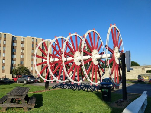 A large, colorful sculpture of six red and white wagon wheels stands in a grassy area near a building and parked cars.