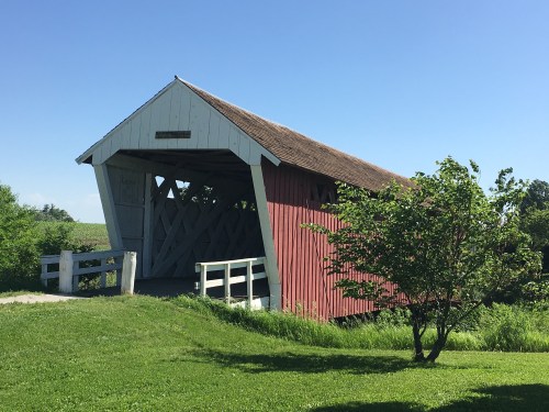 A red and white covered bridge stands in a green landscape under a clear blue sky, surrounded by grass and trees.