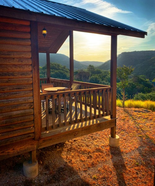A wooden cabin with a porch overlooks a scenic valley at sunset, surrounded by lush hills and a clear sky.