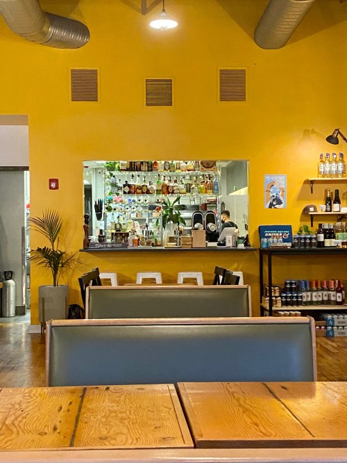 Interior of a vibrant yellow restaurant with seating, a bar area in the background, and shelves of bottles.