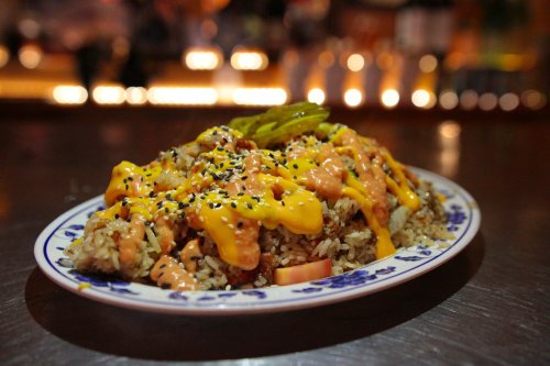 A plate of rice topped with cheese, sauce, pickles, and sesame seeds, served in a vibrant setting.
