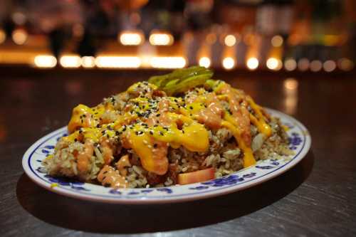 A plate of rice topped with cheese, sauce, pickles, and sesame seeds, served in a vibrant setting.