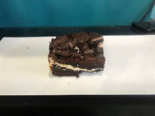 A slice of brownie with a creamy filling, placed on a white plate against a teal background.