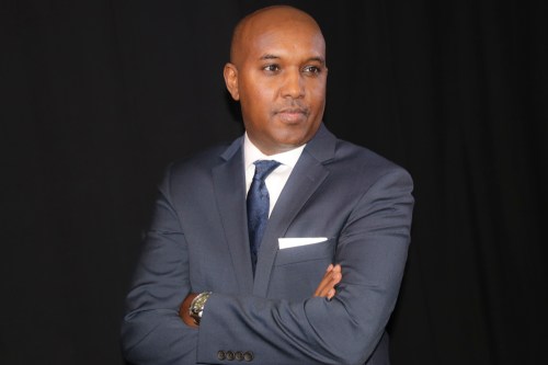 A man in a suit with arms crossed, looking confidently at the camera against a dark background.