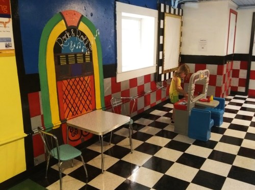 A colorful playroom with a checkered floor, featuring a child playing at a toy kitchen near a vibrant wall mural.
