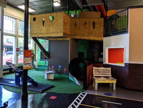Indoor play area featuring a wooden play structure, slide, and colorful decorations with grass-like flooring.