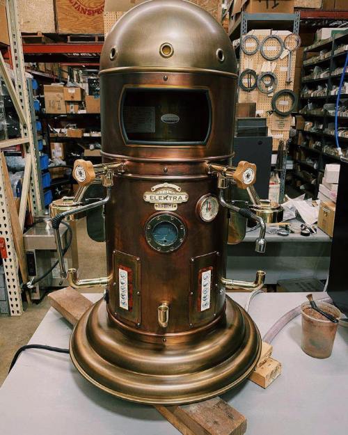 A vintage-style espresso machine with a shiny brass finish, featuring dials and levers, set in a workshop.