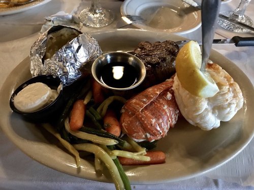 A plate featuring a baked potato, steak, lobster tail, and mixed vegetables with a lemon wedge and dipping sauce.