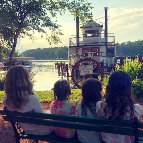 Four children sit on a bench by a lake, watching a steamboat docked nearby under a sunny sky.