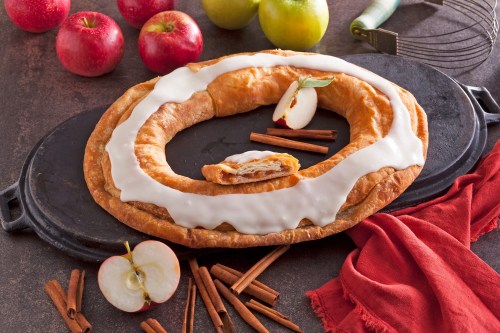 A large, glazed pastry shaped like a ring, surrounded by apples and cinnamon sticks on a dark surface.