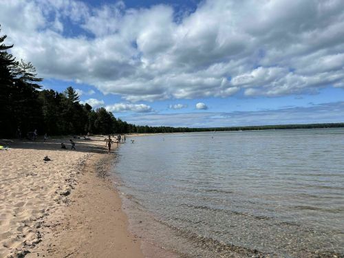 A sandy beach with people enjoying the water, surrounded by trees and a cloudy sky.