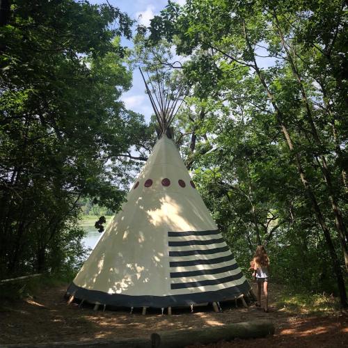 A person stands near a teepee surrounded by lush green trees and a serene lake in the background.
