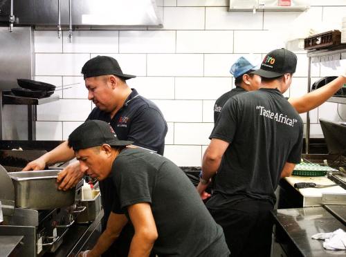 Four chefs in a busy kitchen, wearing black shirts and caps, preparing food and working at various stations.