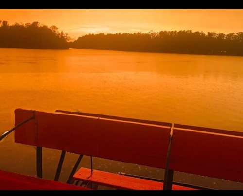 A serene lake at sunset, with vibrant orange hues reflecting on the water and a wooden bench in the foreground.