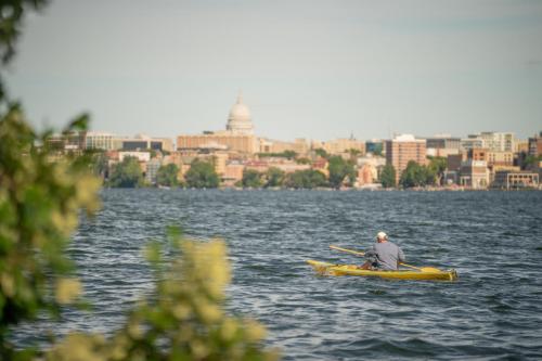 A person kayaking on a lake with a city skyline and dome building in the background, framed by greenery.