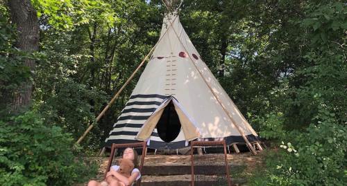 A teepee nestled in a wooded area, with a person lounging in a chair nearby, surrounded by greenery.