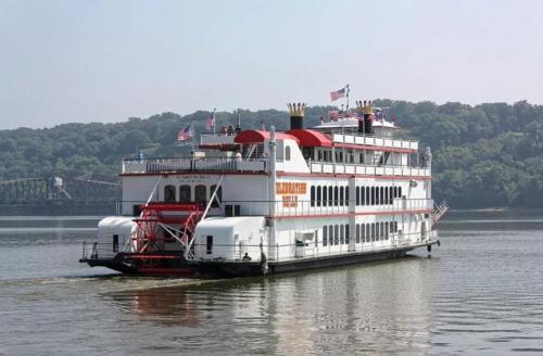 A large white riverboat with red accents and flags cruising on a calm river, surrounded by green trees.