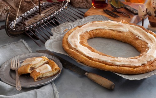 A large circular pastry with cream frosting, alongside a slice on a plate, with decorative items in the background.