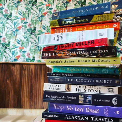 A stack of books on a floral wallpaper background, featuring titles like "Death in Florence" and "Angela's Ashes."