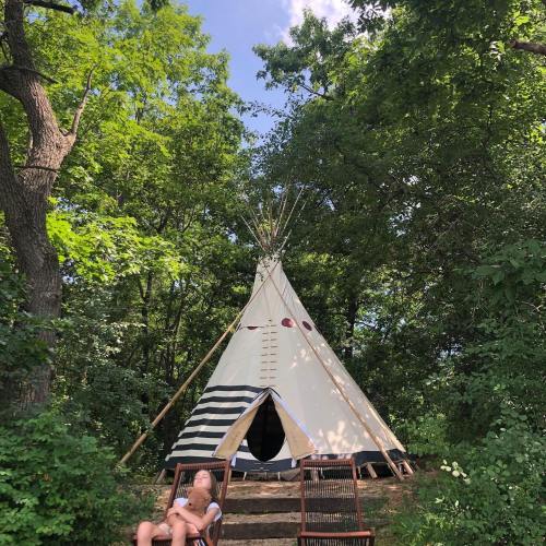 A teepee nestled in a lush green forest, with a person relaxing in a chair nearby.