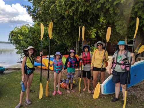A group of five people in life jackets and hats, holding paddles, standing by a lake with kayaks in the background.