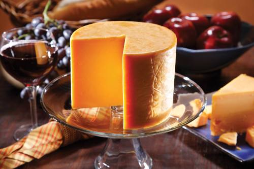 A round cheese wheel on a glass stand, surrounded by wine, grapes, apples, and slices of cheese on a plate.