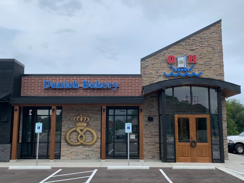 Exterior of a Danish Bakery with a stone facade, blue signage, and decorative elements, set against a cloudy sky.