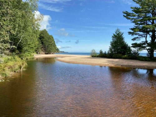 A serene landscape featuring a calm river flowing into a sandy beach, surrounded by lush green trees and a clear blue sky.