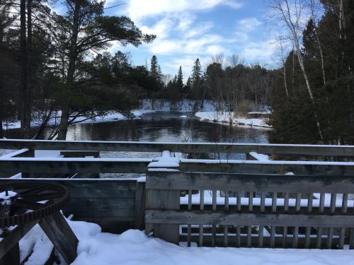 A snowy landscape with a river surrounded by trees, viewed from a wooden deck.