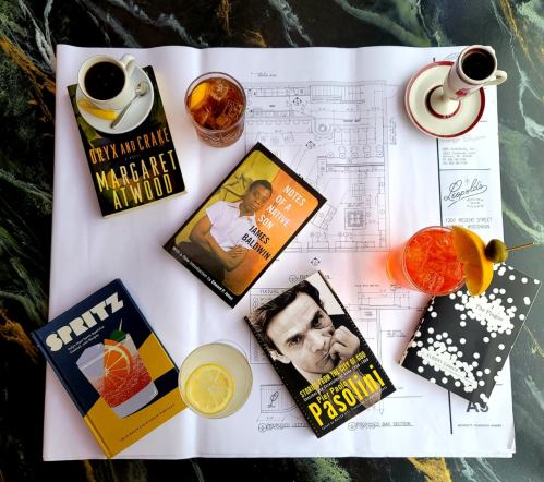 A flat lay of drinks, books, and a blueprint on a marble surface, featuring titles by Atwood and Pasolini.