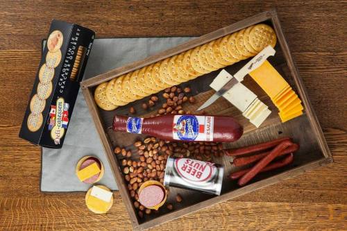 A wooden tray filled with crackers, cheese slices, sausages, nuts, and a bottle of sauce, arranged neatly.