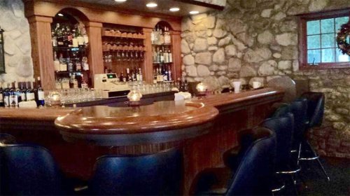A cozy bar with a wooden counter, stone walls, and shelves lined with various bottles of liquor.