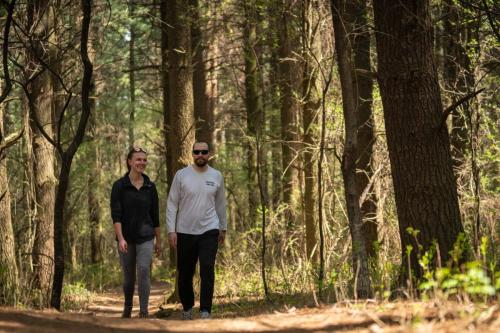 Two people walking on a forest trail, surrounded by tall trees and greenery, enjoying a sunny day outdoors.