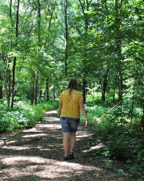 A person walks along a wooded path, surrounded by lush green trees and foliage.