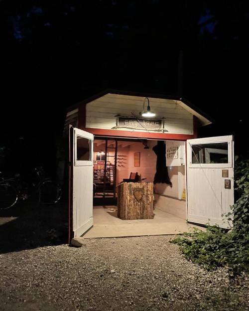 A small, illuminated shed at night with open doors, featuring a wooden table inside and surrounded by greenery.