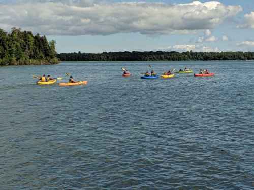 A group of kayakers in colorful kayaks paddling on a calm lake surrounded by trees and a blue sky.