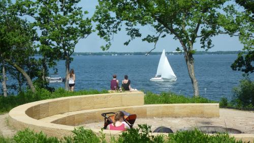 Three people sit on a curved stone bench by a lake, watching a sailboat glide across the water on a sunny day.