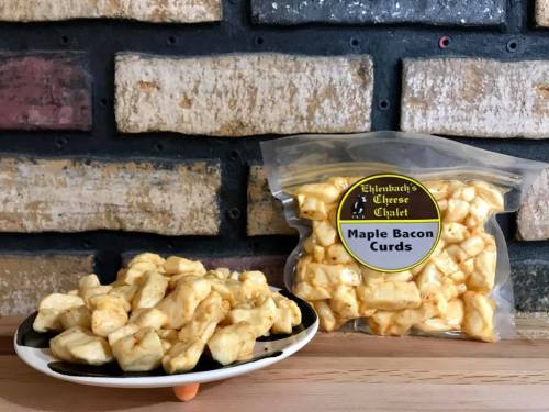 A plate of cheese curds next to a bag labeled "Maple Bacon Curds" against a brick wall background.