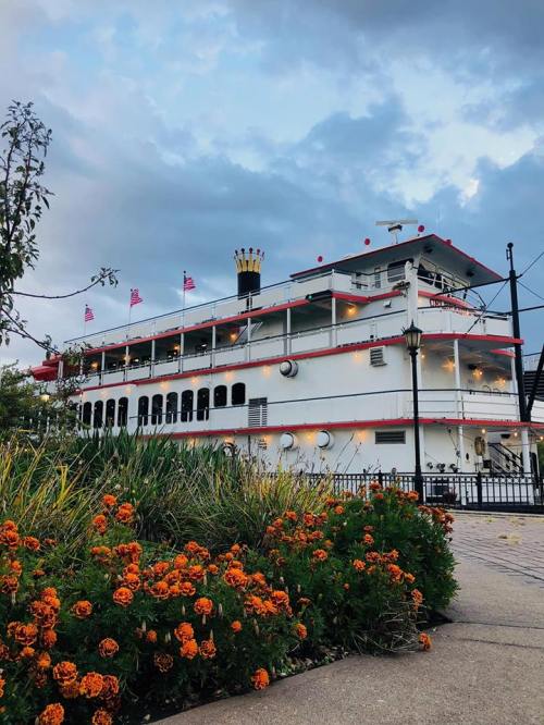 A large riverboat with red and white details, surrounded by colorful flowers and under a cloudy sky.
