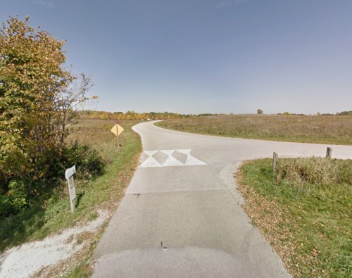 A winding road with a warning sign and a speed bump, surrounded by open fields and trees in the background.