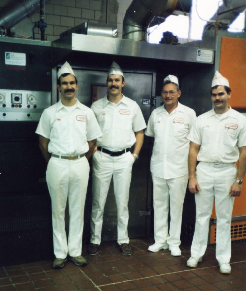 Four men in white uniforms and paper hats stand in front of industrial equipment, smiling at the camera.
