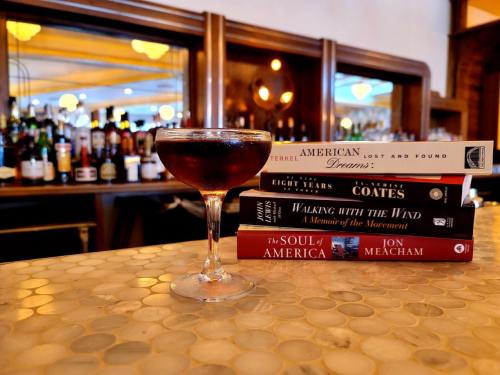 A cocktail sits on a bar counter next to a stack of books, with a blurred bar backdrop.