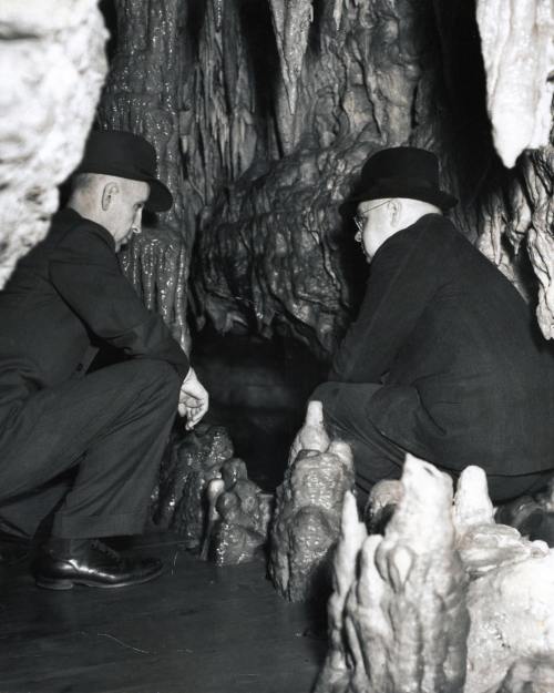 Two men in hats examine a cave formation, surrounded by stalactites and stalagmites in a dimly lit cavern.