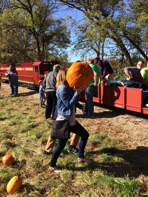 A person carries a large pumpkin near a train, with people and trees in the background on a sunny day.