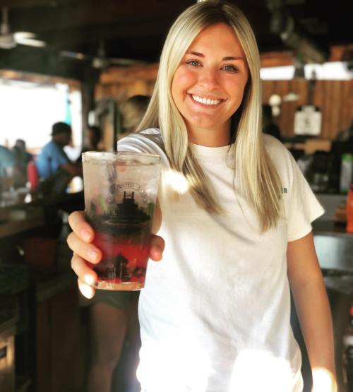 A smiling young woman in a white shirt holds up a drink in a lively bar setting.
